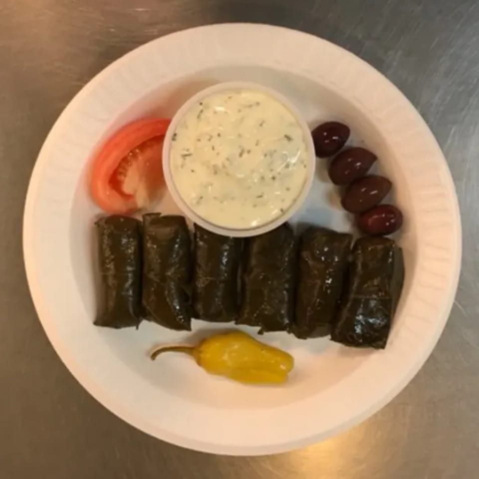 A Greek appetizer plate with dolmades, tzatziki, tomato, olives, and a yellow pepper.